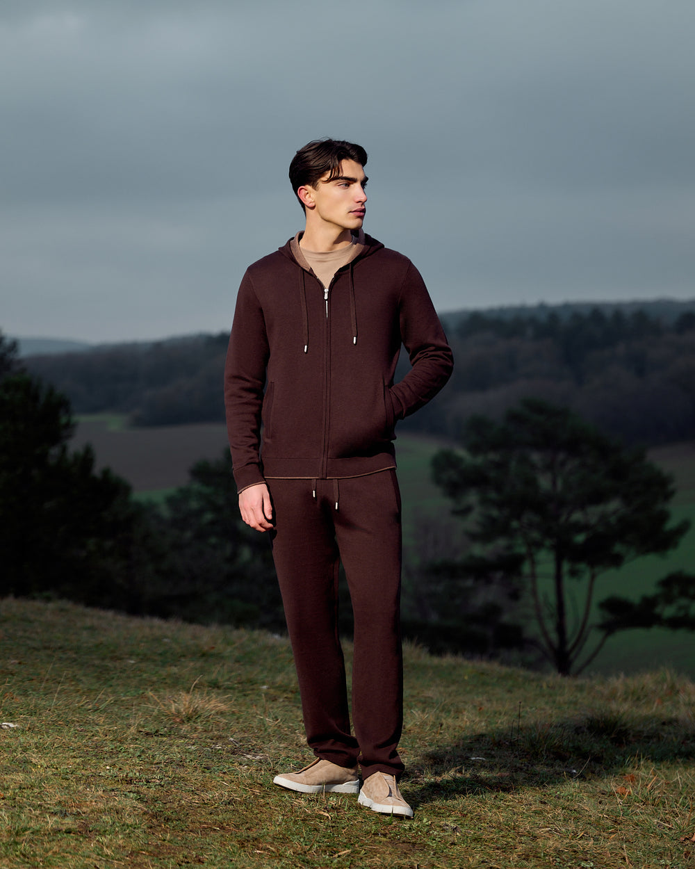 Man wearing a brown tracksuit standing on a grassy hill with a scenic background