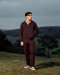 Man wearing a brown tracksuit standing on a grassy hill with a scenic background