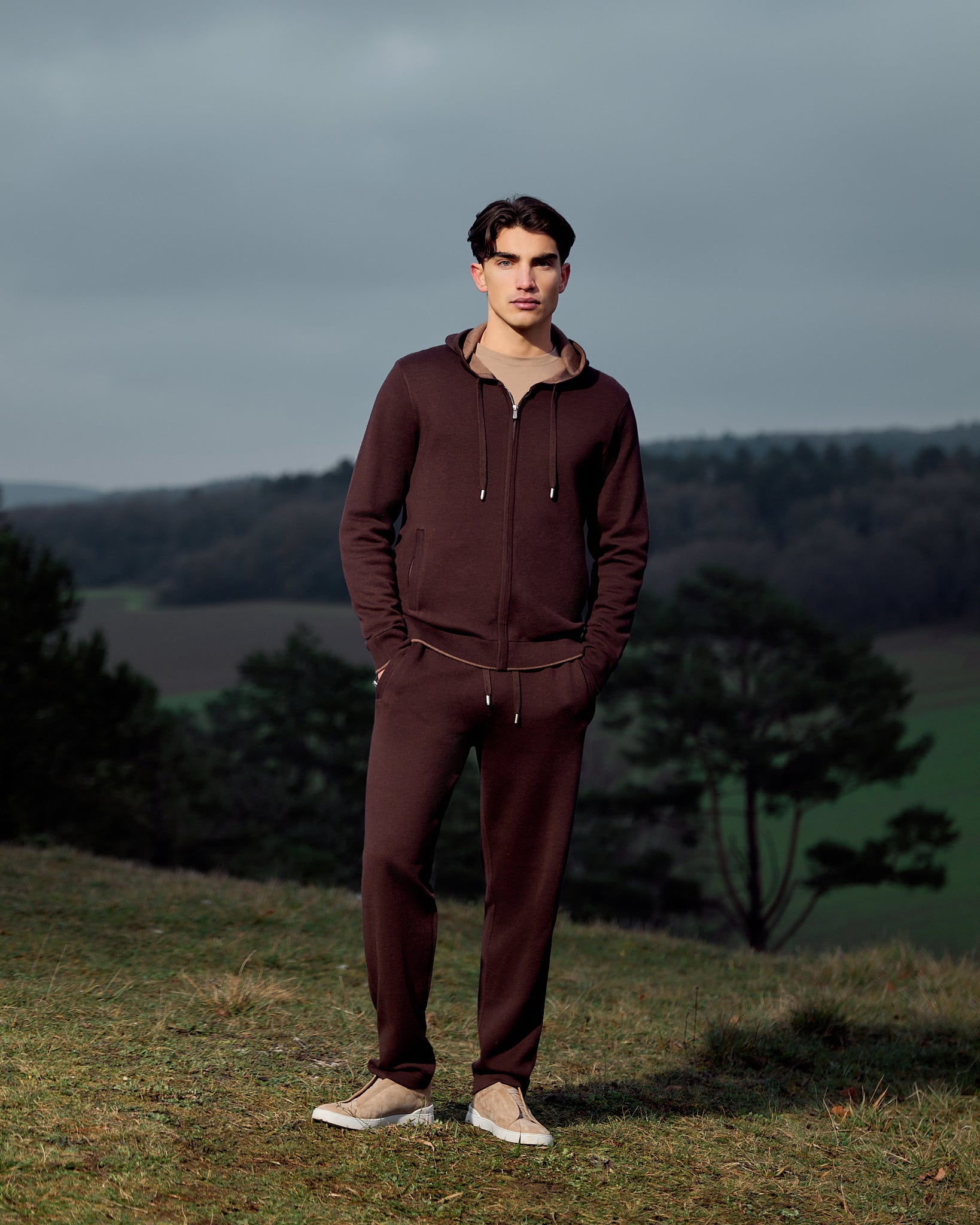 Man wearing a maroon tracksuit standing in a field with trees and a cloudy sky.