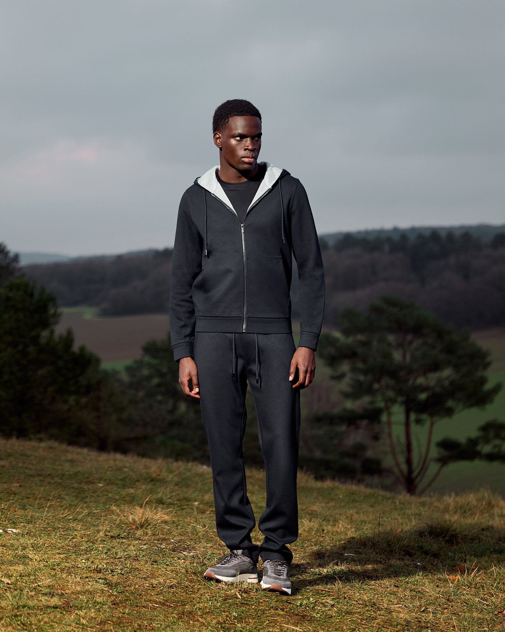 Man wearing a dark gray hoodie and pants standing in a field with trees and a cloudy sky in the background.