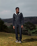 Man wearing a dark gray hoodie and pants standing in a field with trees and a cloudy sky in the background.