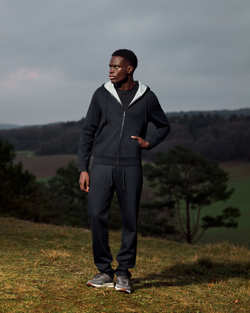 Man wearing a dark hoodie and pants standing in a field with trees and a cloudy sky in the background