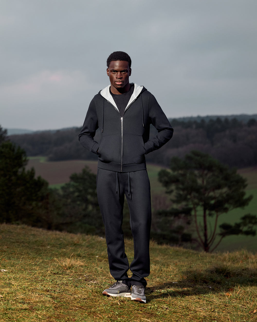 Man wearing a dark gray tracksuit standing on a grassy hill with trees and a cloudy sky in the background