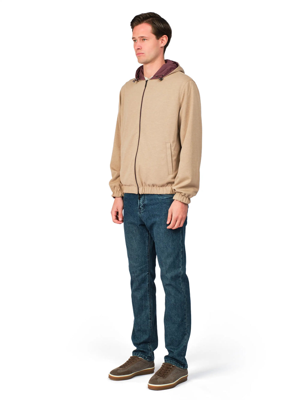 Man wearing a beige jacket with a maroon hood and blue jeans on a white background