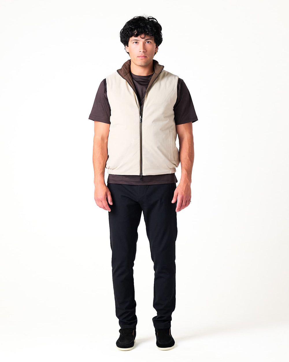 Person wearing a beige vest over a dark shirt with black pants on a white background