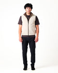 Person wearing a beige vest over a dark shirt with black pants on a white background