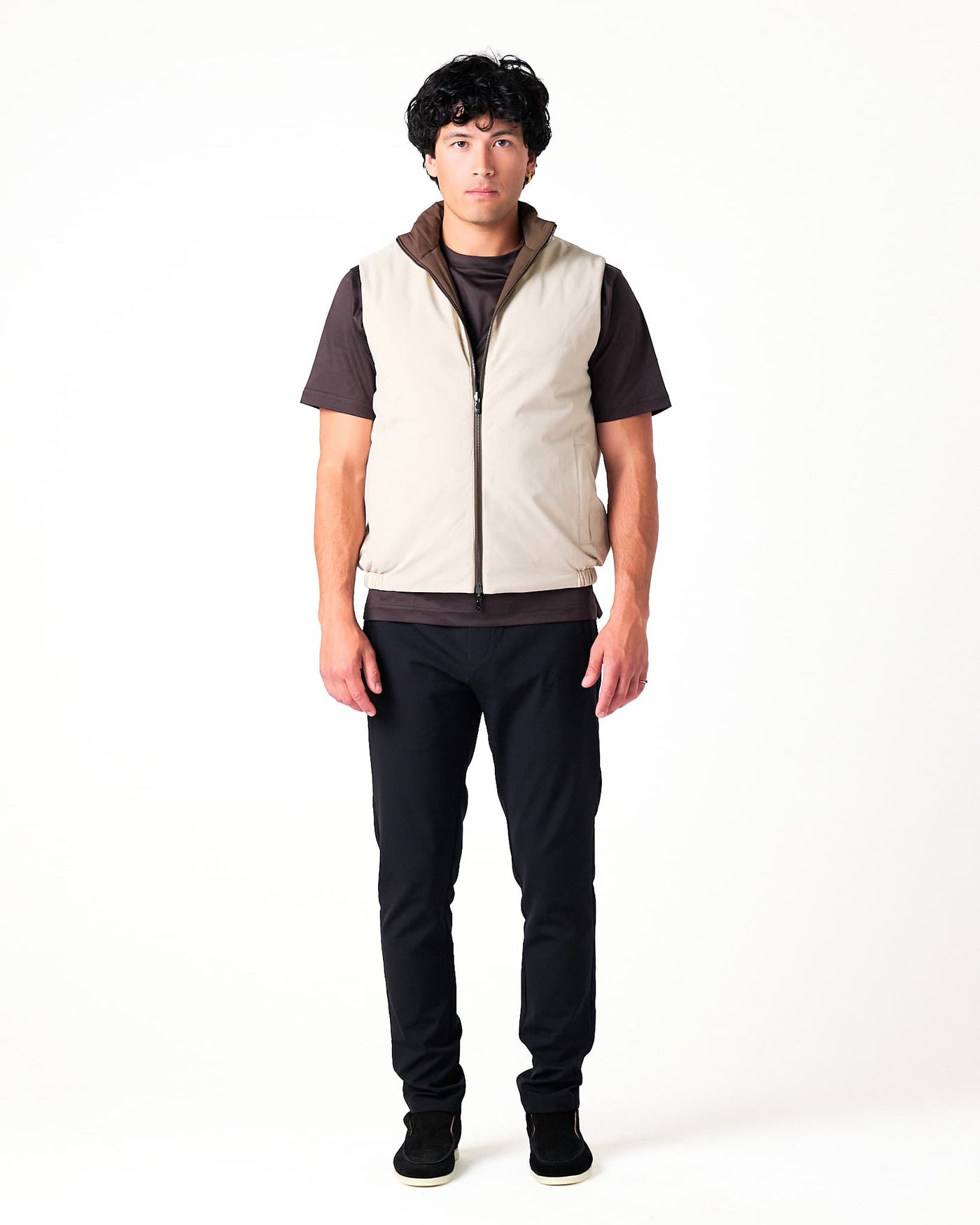 Person wearing a beige vest over a dark shirt with black pants on a white background