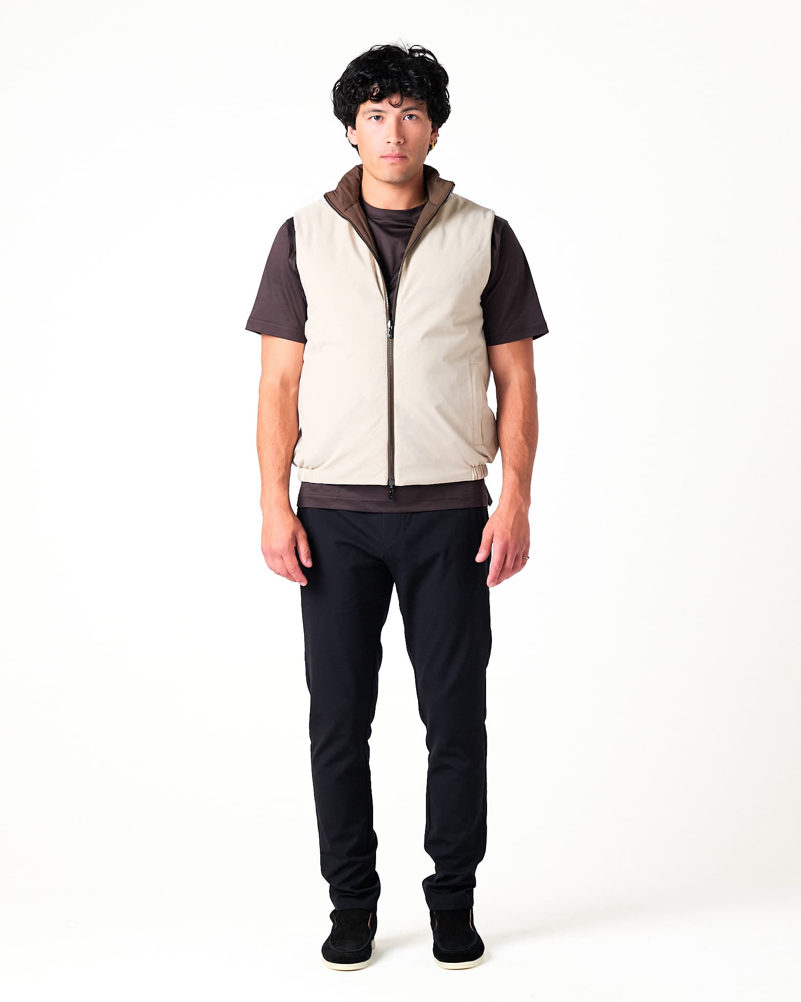 Person wearing a beige vest over a dark shirt with black pants on a white background