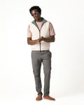Man wearing a light-colored vest over a pink shirt, gray pants, and brown shoes on a white background
