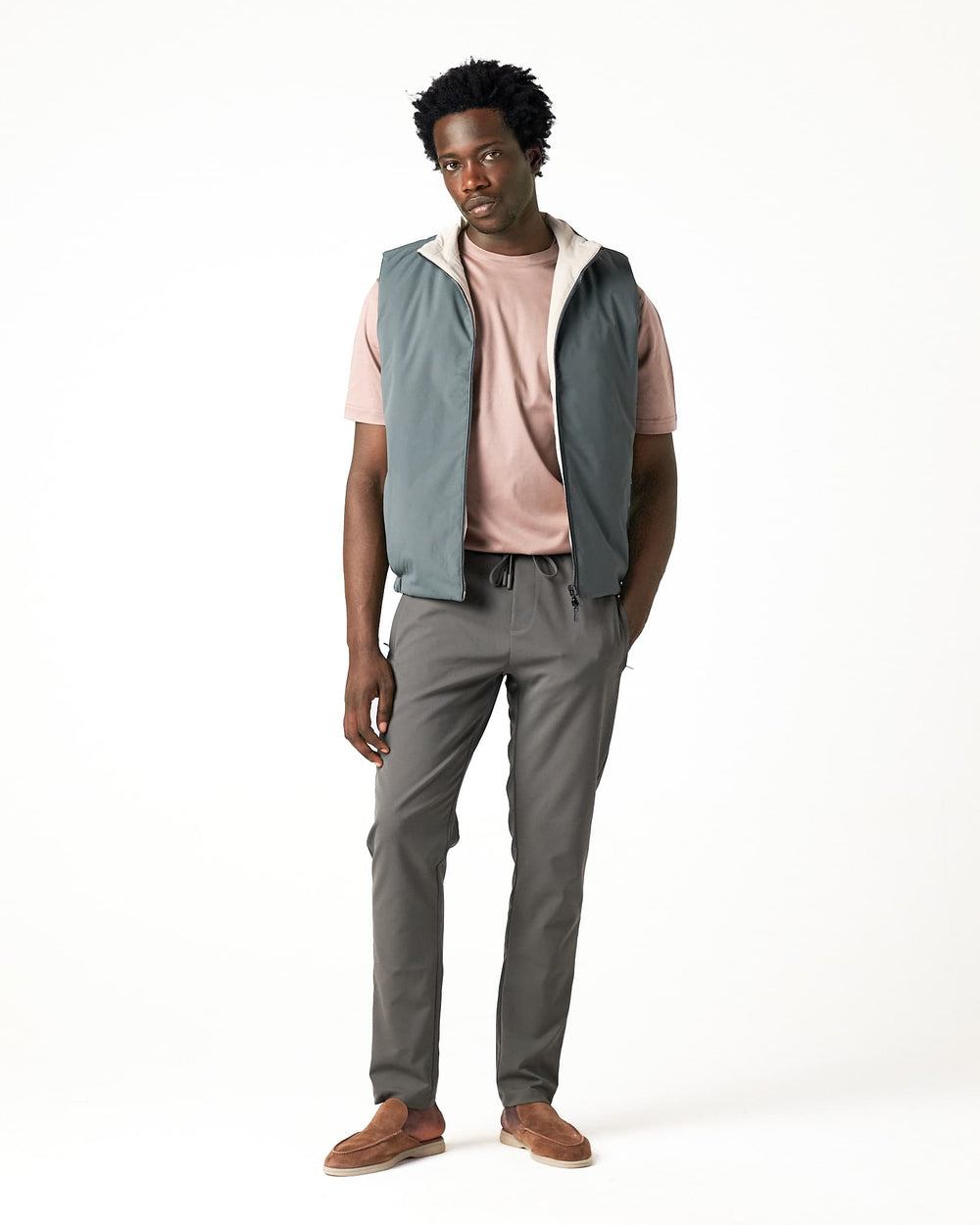 Man wearing a light gray vest over a pink shirt with gray pants on a white background