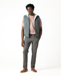 Man wearing a light gray vest over a pink shirt with gray pants on a white background