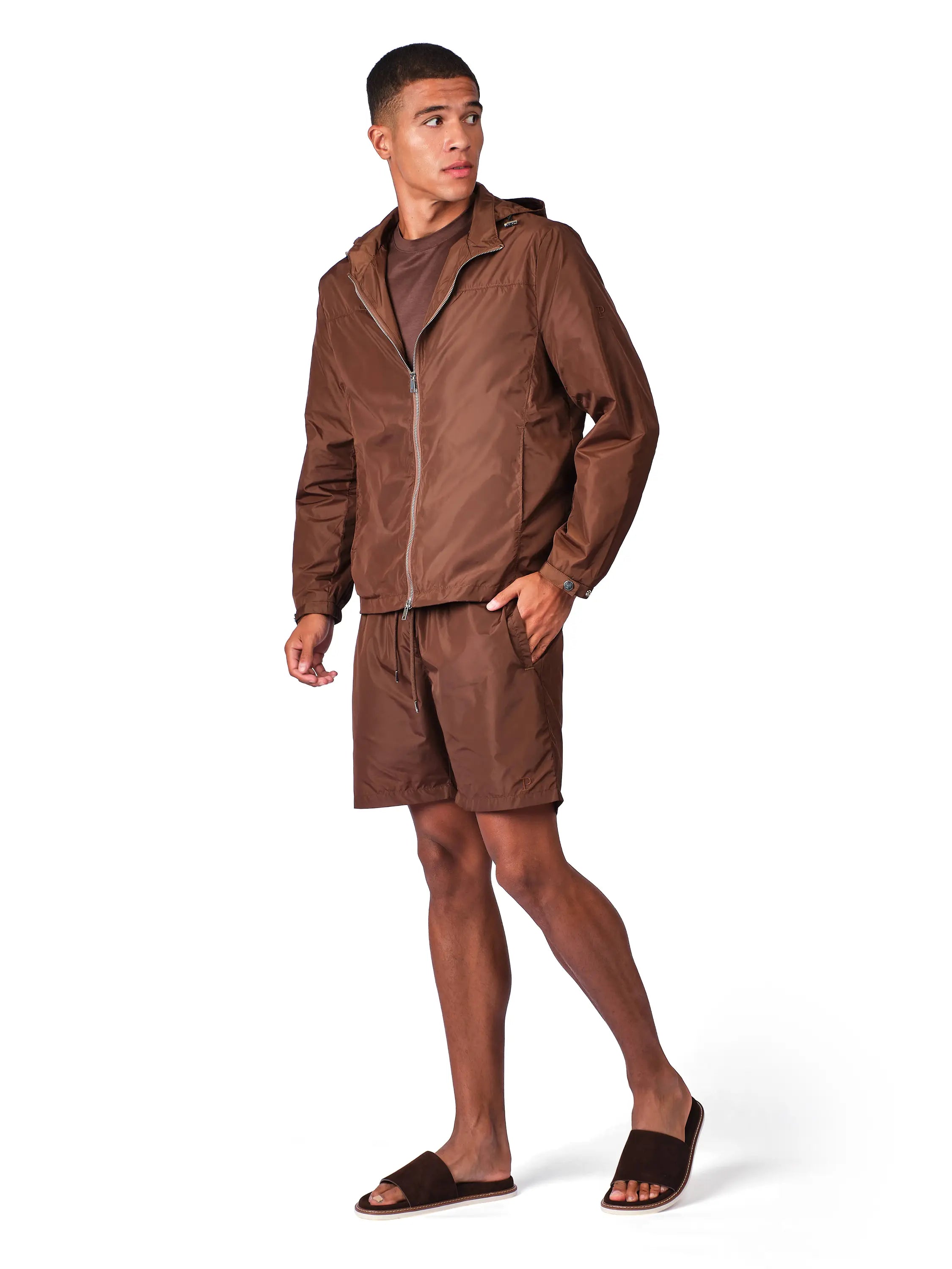 A man modeling a matching brown zip-up hooded jacket and shorts set, paired with a brown shirt and dark brown slide sandals, against a plain white background.