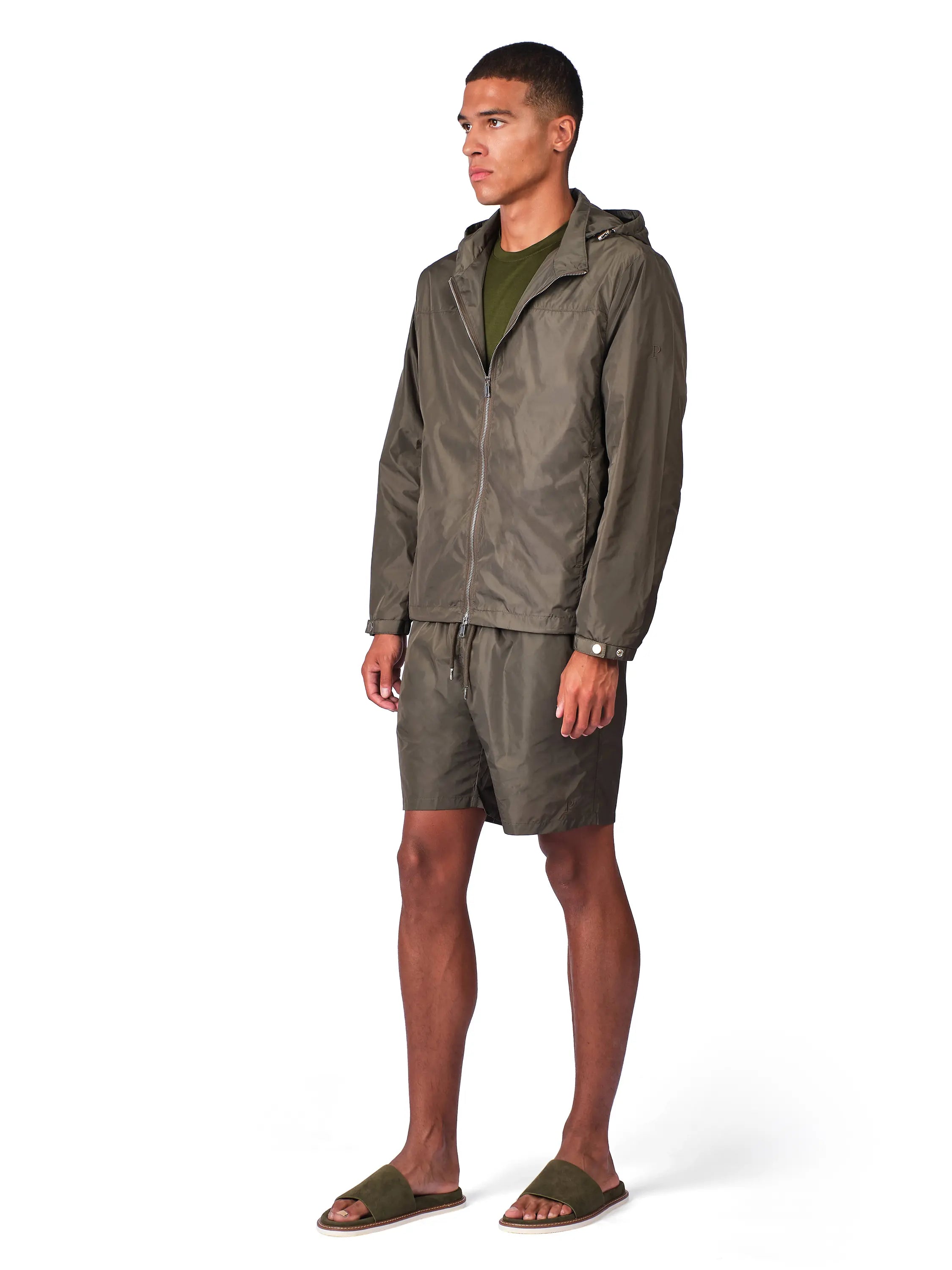 A man modeling a matching olive green outdoor set consisting of a lightweight zip-up hooded jacket and coordinating shorts, worn over an olive green t-shirt and paired with brown slide sandals, on a white studio background.