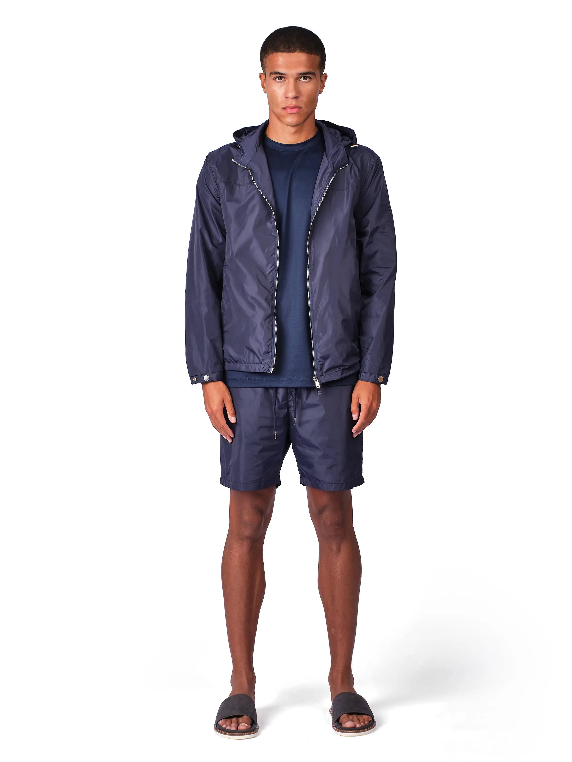 A man modeling a matching navy blue lightweight hooded jacket and shorts set, paired with a navy t-shirt and dark slide sandals, against a plain white background.