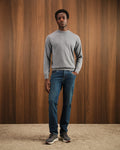 Man wearing a gray sweater and blue jeans standing against a wooden wall.