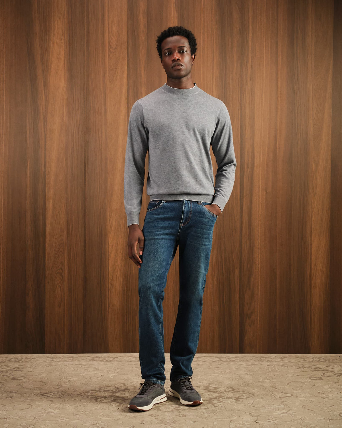 Man wearing a gray sweater and blue jeans standing against a wooden wall.