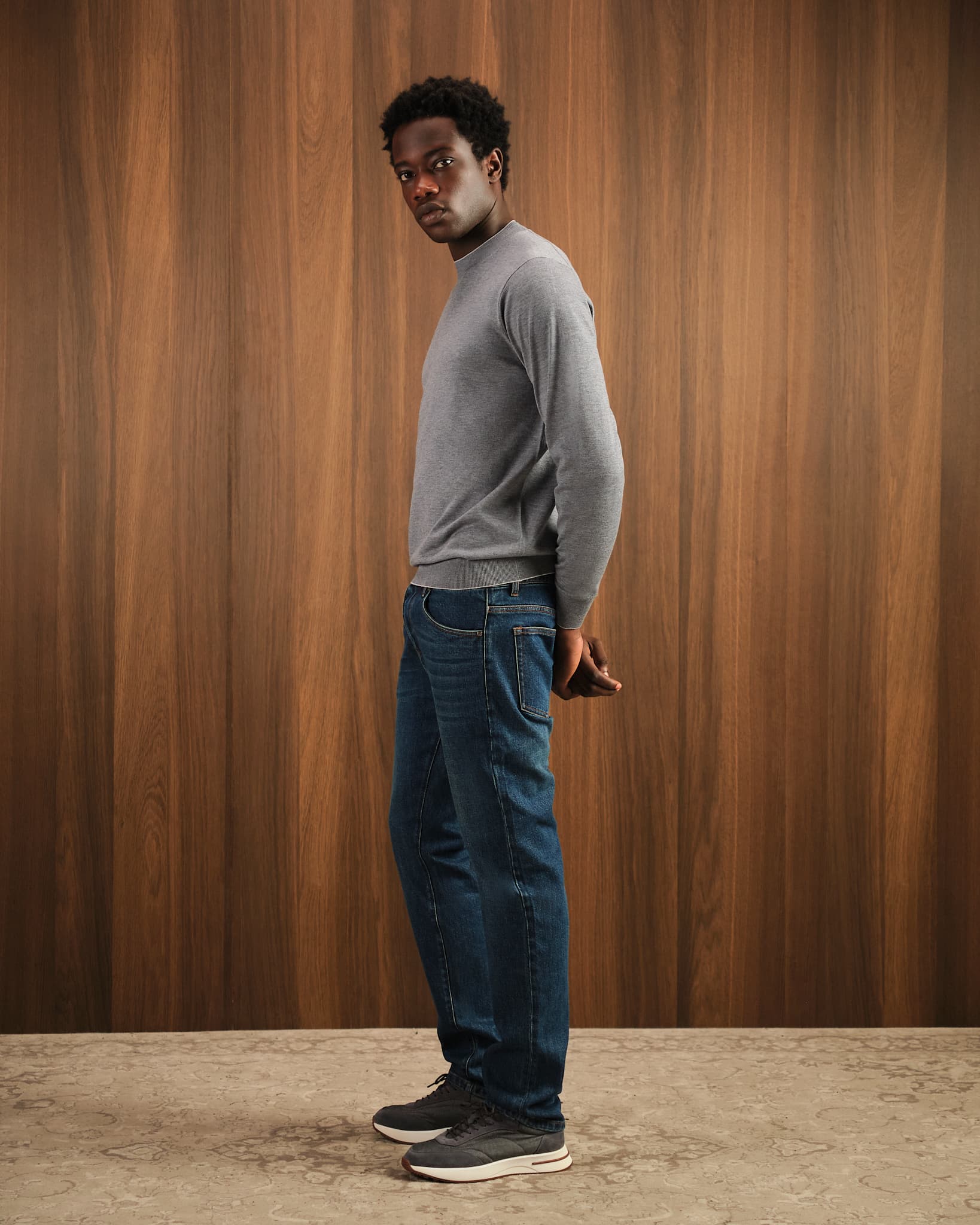 Man wearing a gray sweater and blue jeans standing against a wooden paneled wall.