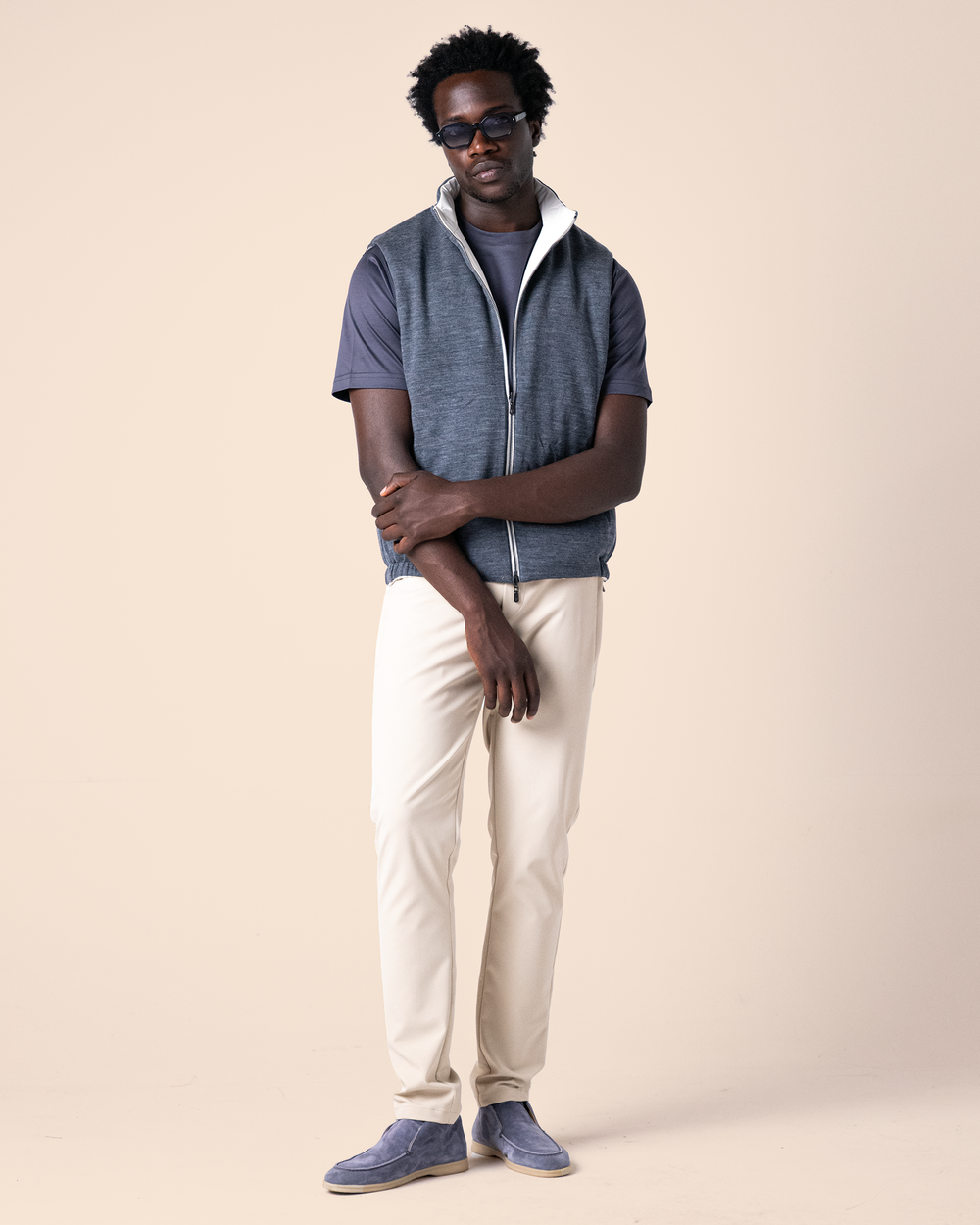 Man wearing a blue vest over a gray shirt, white pants, and blue shoes on a beige background