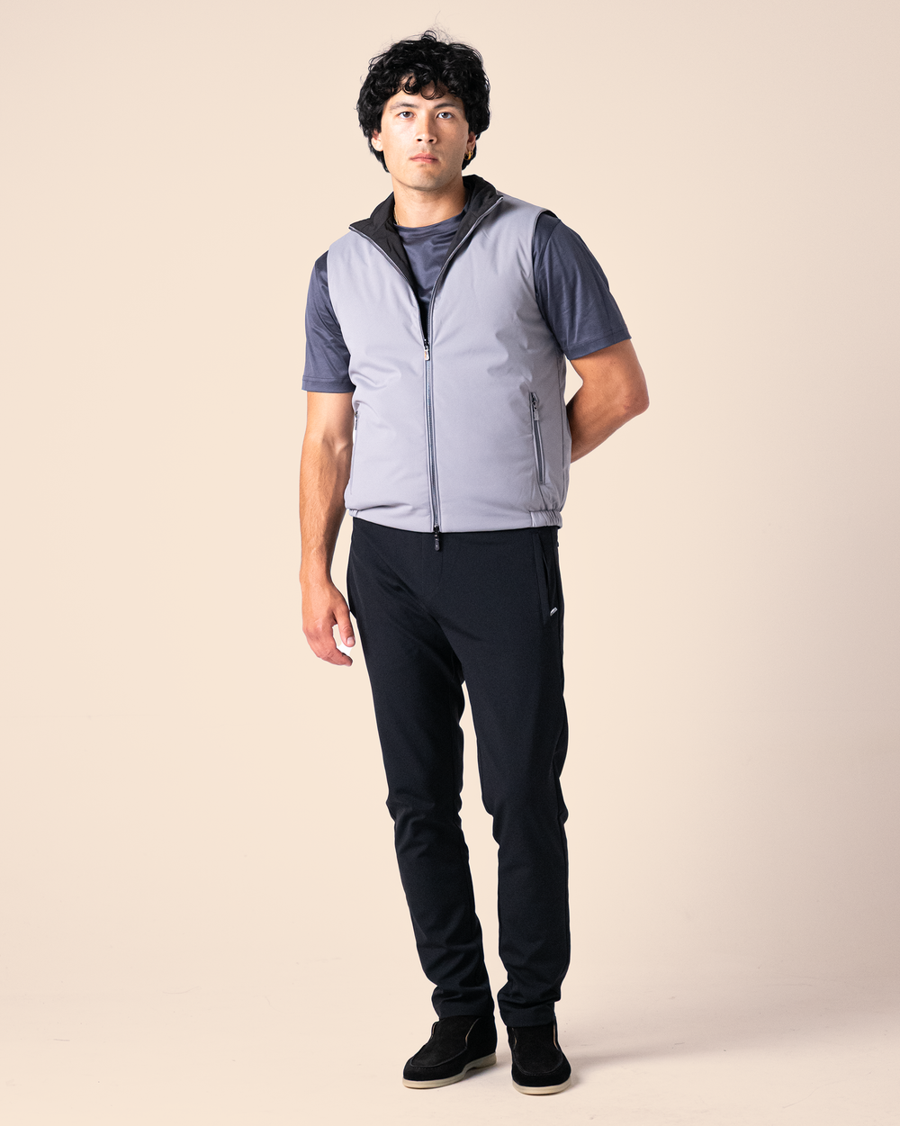 Man wearing a gray vest over a dark shirt with black pants on a beige background