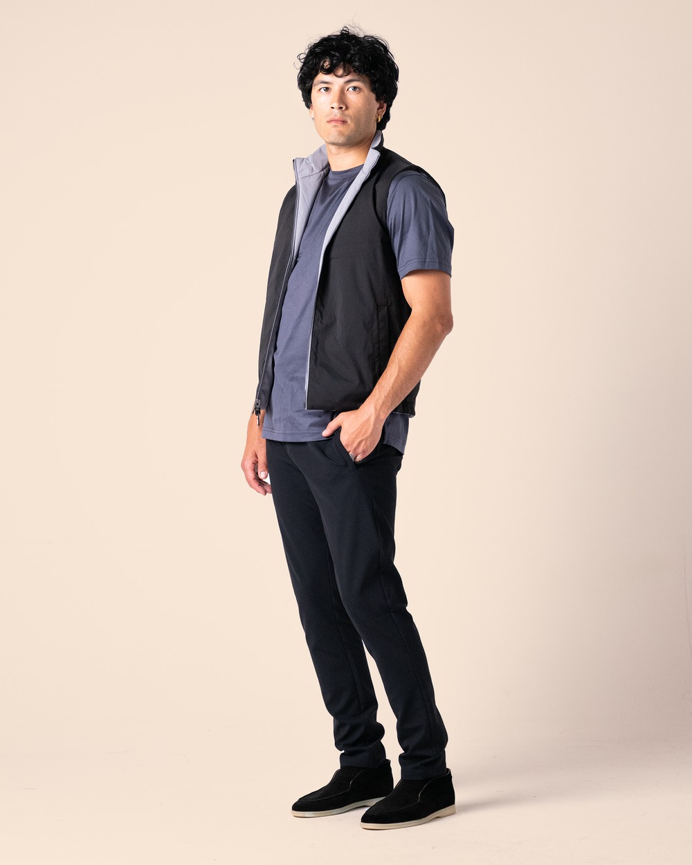 Man wearing a dark vest over a blue shirt with black pants on a beige background