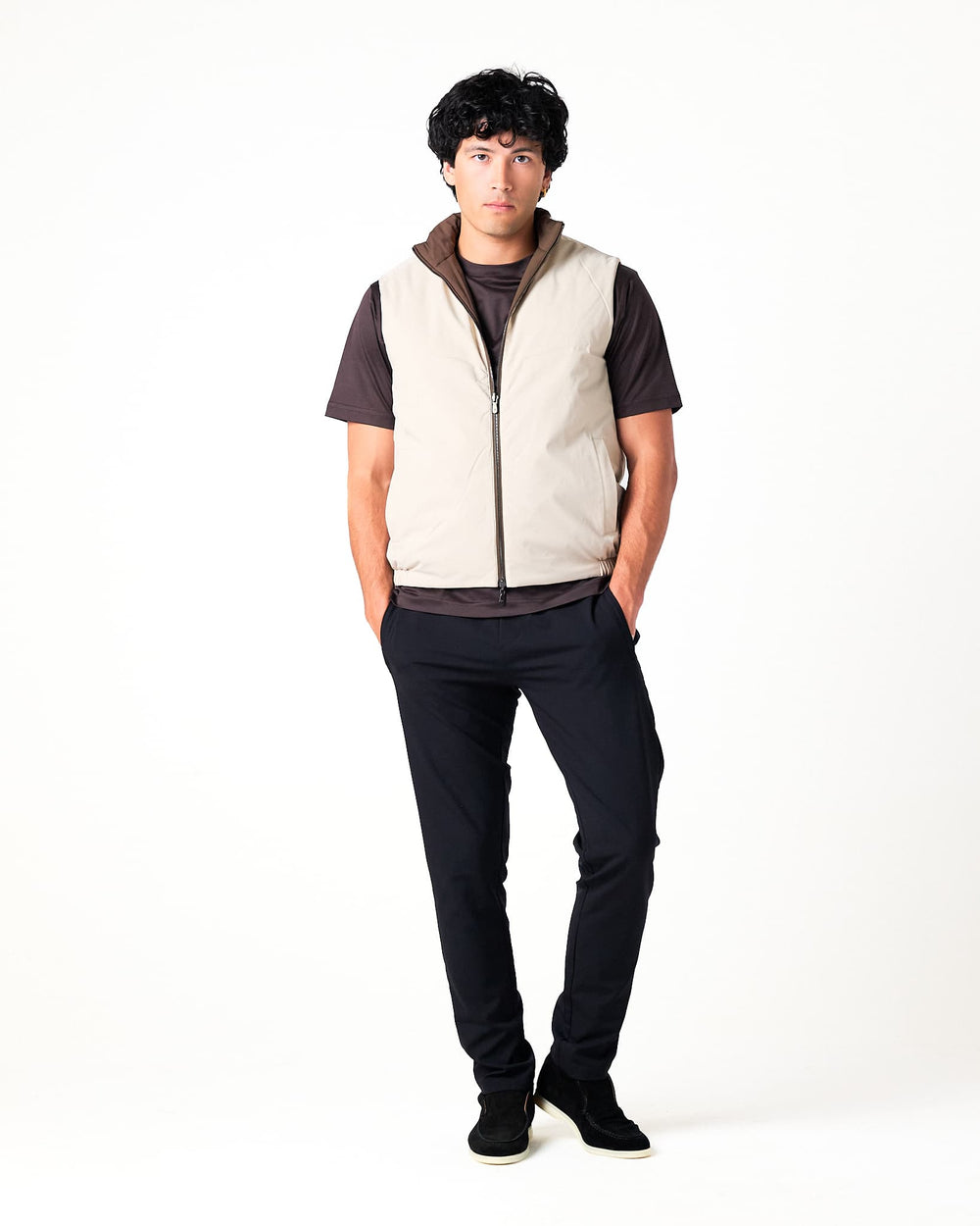 Person wearing a beige vest over a dark shirt with black pants on a white background