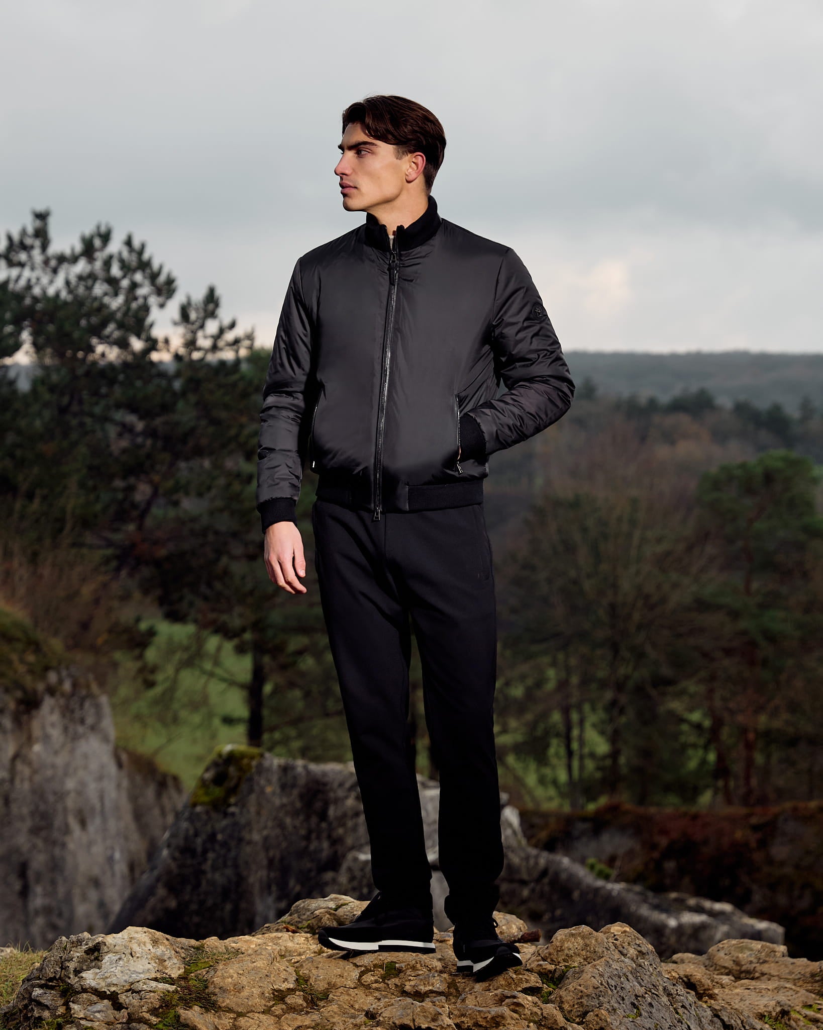 Man in a black outfit standing on a rocky outcrop with a forest background