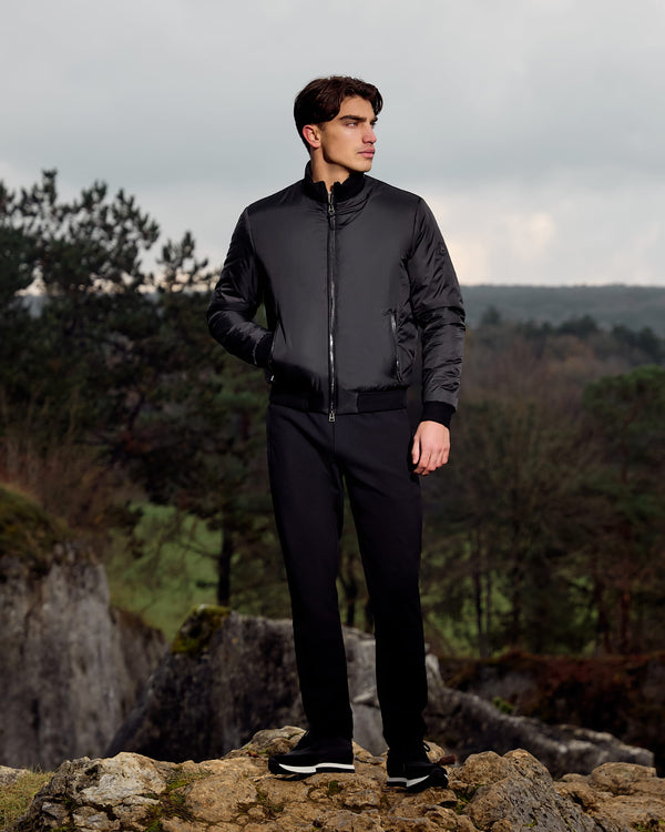 Man wearing a black jacket and pants standing on a rocky outcrop with a forest background