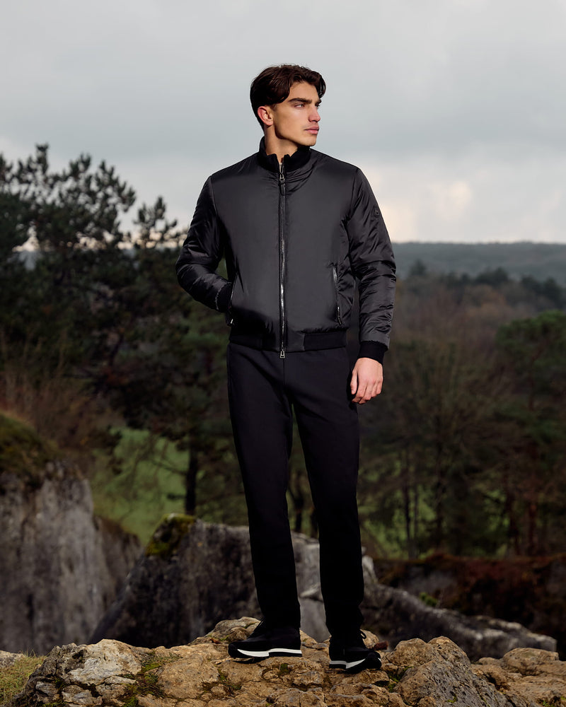 Man wearing a black jacket and pants standing on a rocky outcrop with a forest background