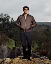 Man wearing a brown jacket and black pants standing on a rocky outcrop with a forest background