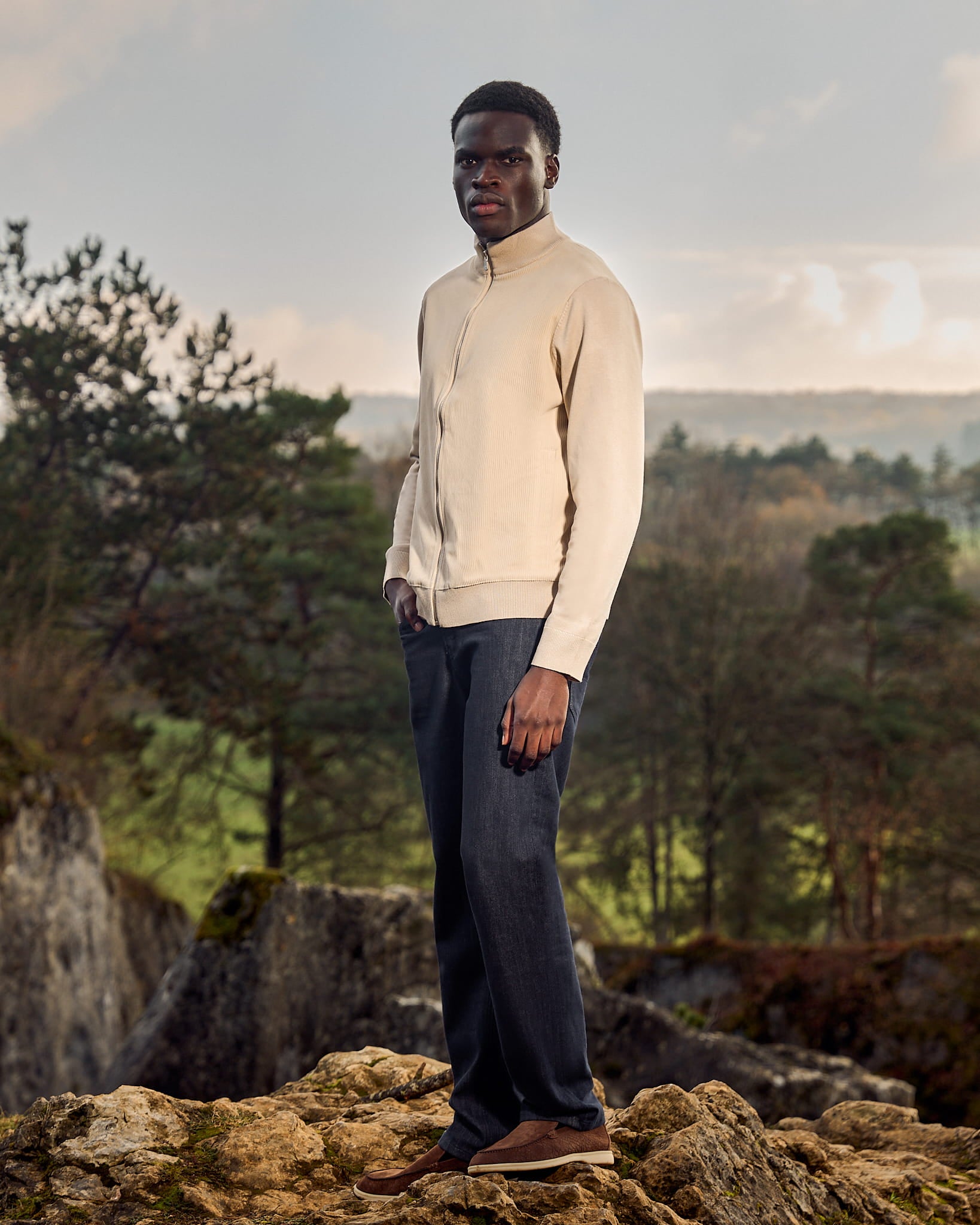 Man standing on a rocky outcrop with a scenic background of trees and sky.
