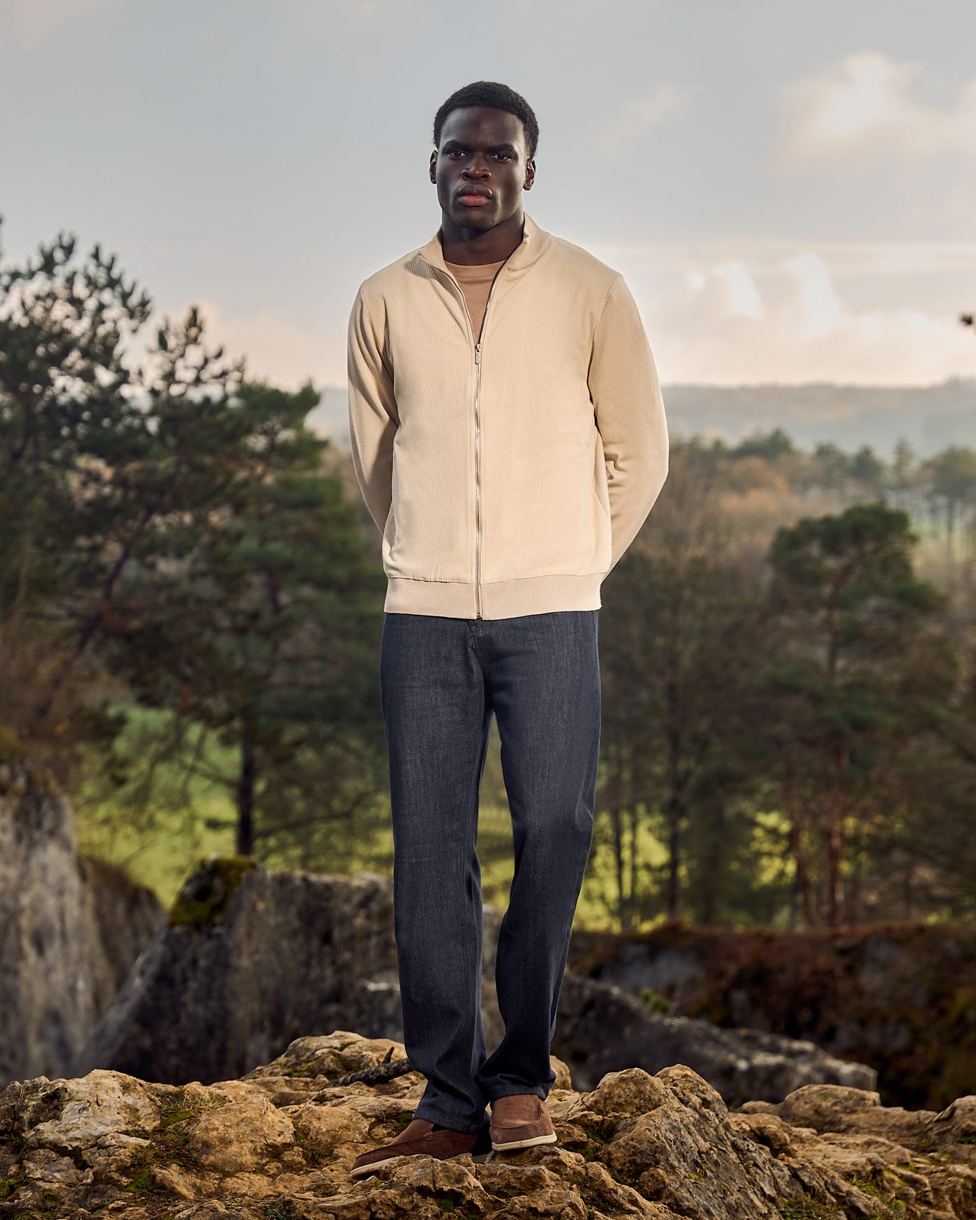Man standing on a rocky outcrop with a scenic background of trees and mountains.