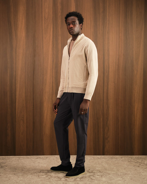 Man wearing a beige sweater and dark pants against a wooden paneled wall