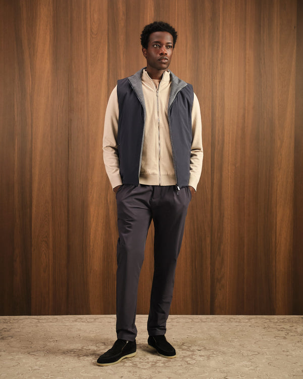 Man wearing a beige shirt, dark vest, and gray pants against a wooden paneled wall.