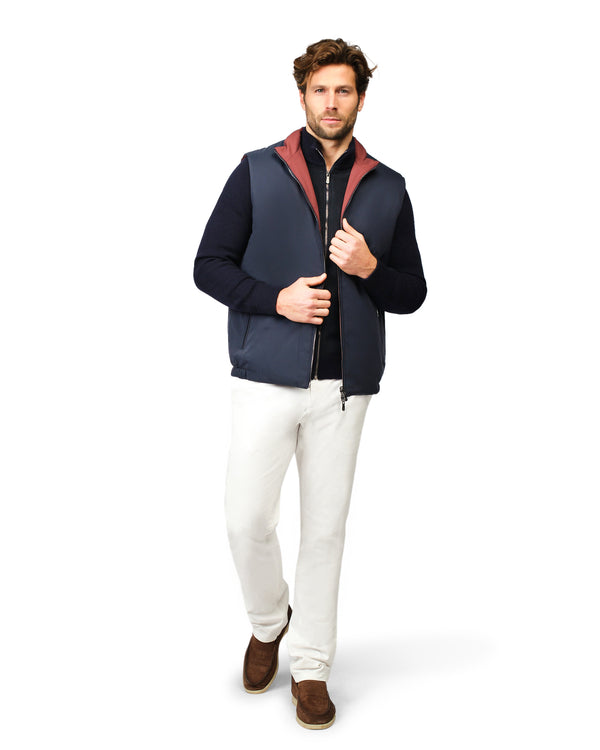 Man wearing a navy blue vest over a black shirt with white pants on a white background