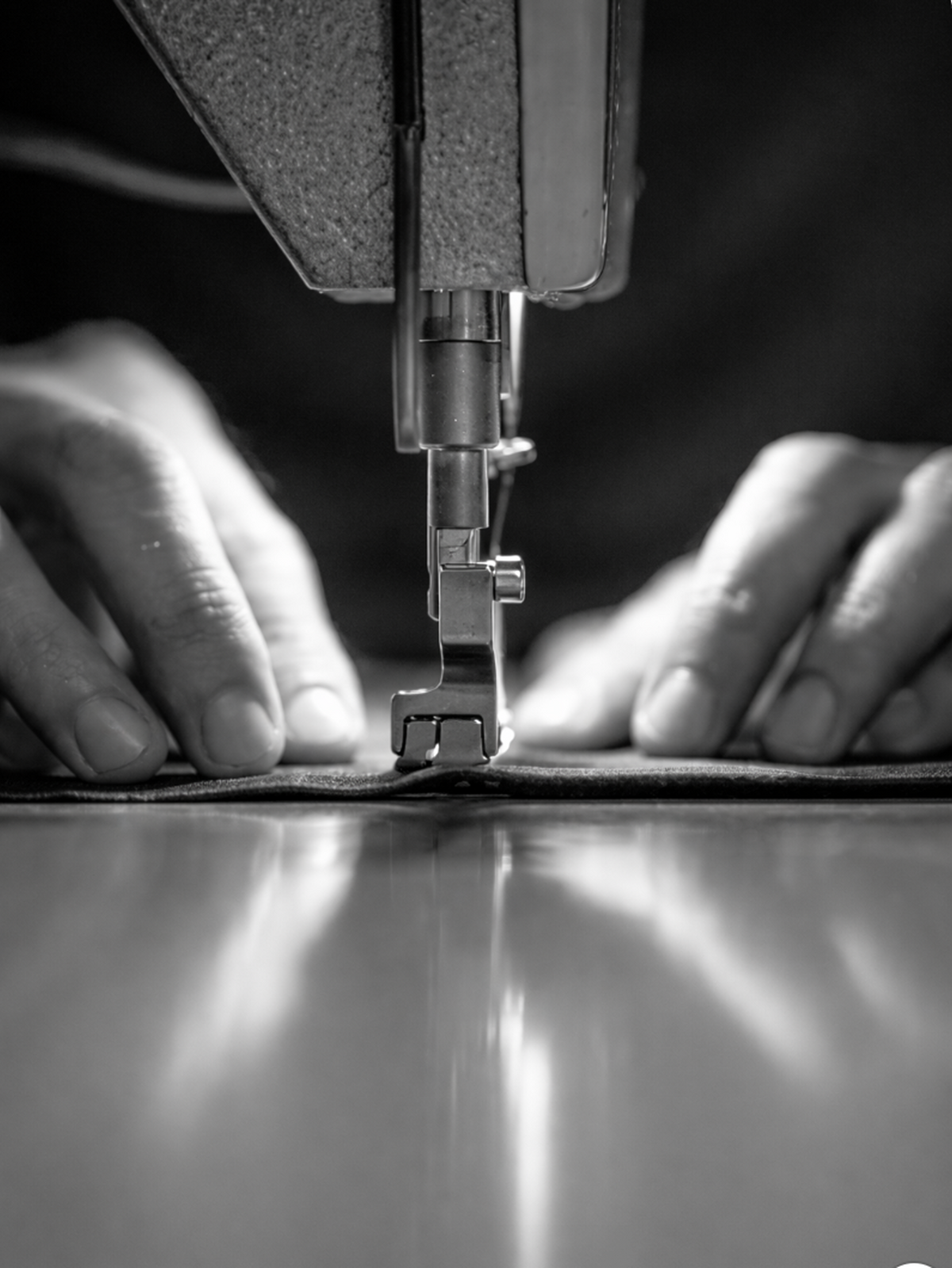 Close-up of a sewing machine needle piercing fabric with hands guiding it, in black and white.