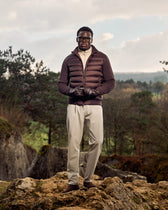 Man wearing a brown puffer jacket and beige pants standing on rocky terrain with trees and sky in the background.