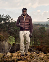 Man wearing a brown puffer jacket and sunglasses standing on a rocky outcrop with trees and sky in the background.