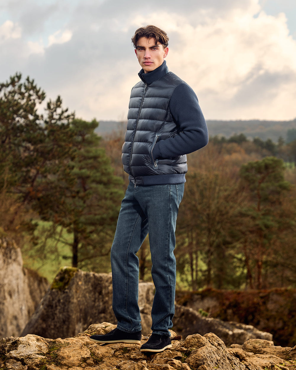 Man wearing a blue puffer jacket and jeans standing on rocky terrain with trees in the background
