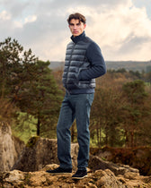 Man wearing a blue puffer jacket and jeans standing on rocky terrain with trees in the background