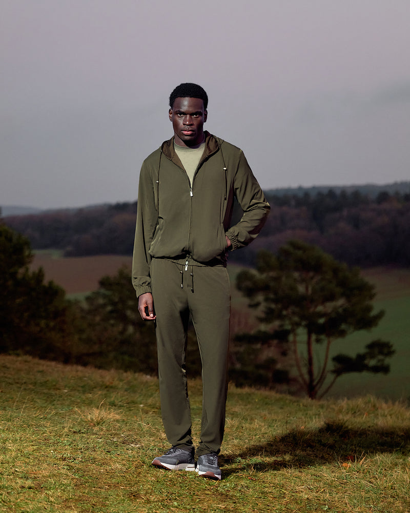 Man wearing a green tracksuit standing in a field with trees and a cloudy sky in the background