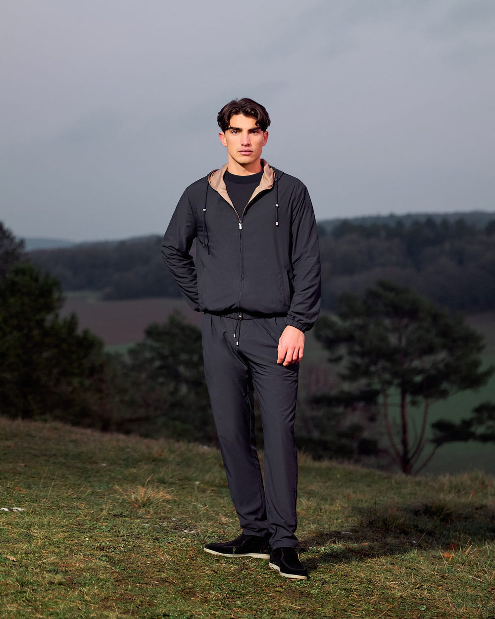 Man wearing a dark jacket and pants standing in a natural setting with trees and grass.
