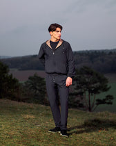 Man wearing a dark jacket and pants standing in a natural landscape with trees and open sky.