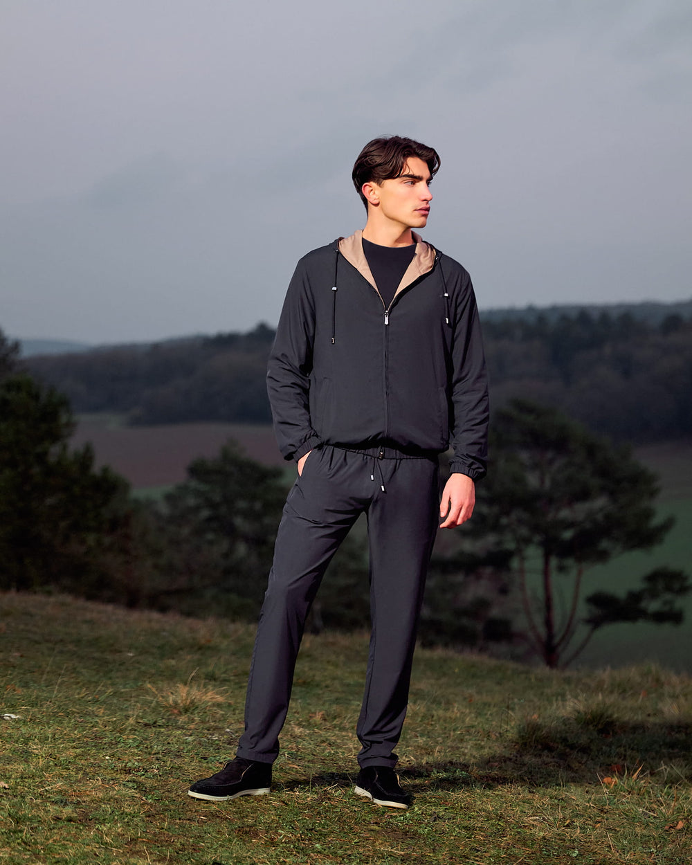 Man wearing a dark gray tracksuit standing on a grassy hill with trees and a cloudy sky in the background.