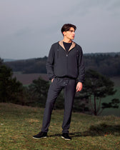 Man wearing a dark gray tracksuit standing on a grassy hill with trees and a cloudy sky in the background.