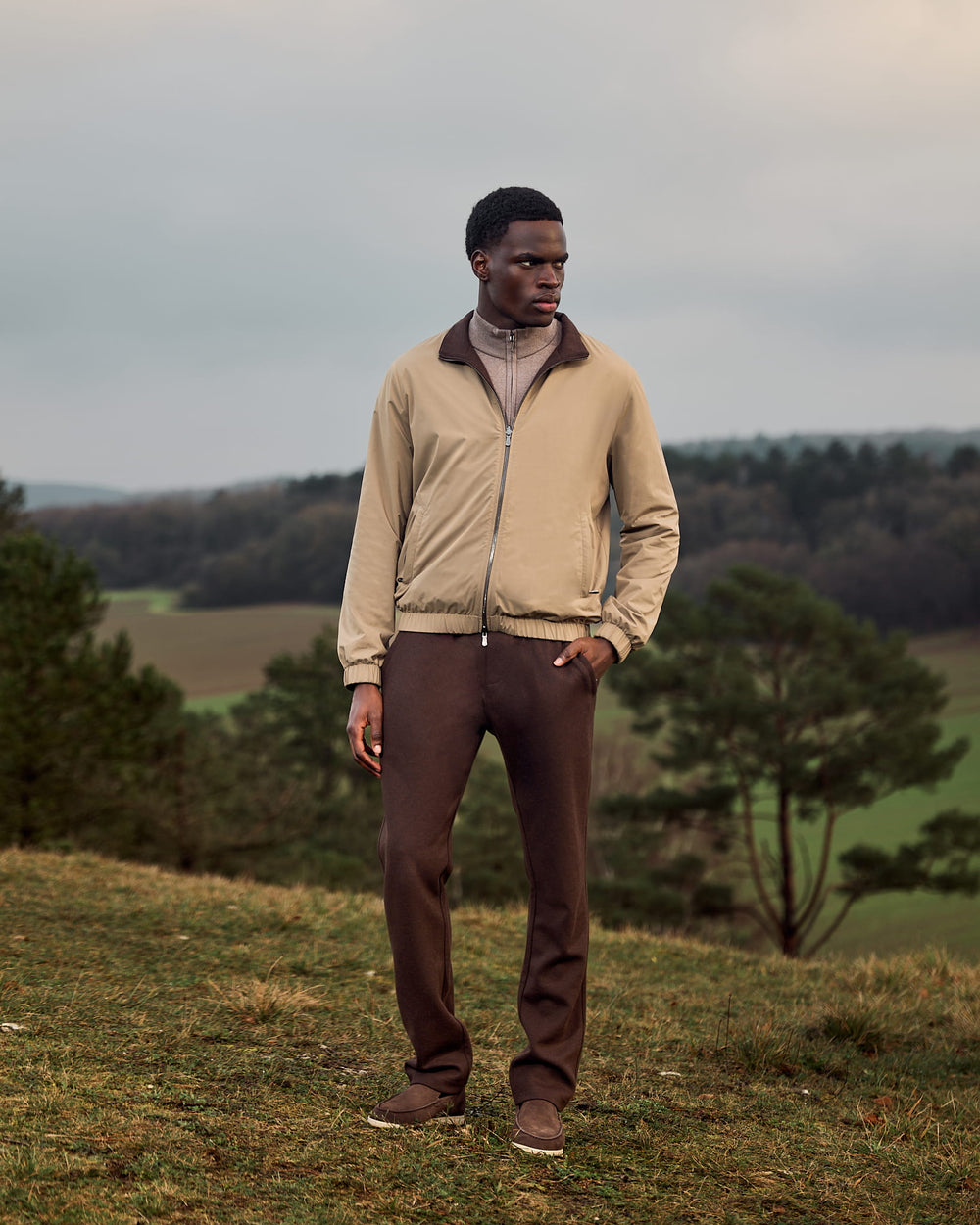 Man standing in a field wearing a beige jacket and brown pants.