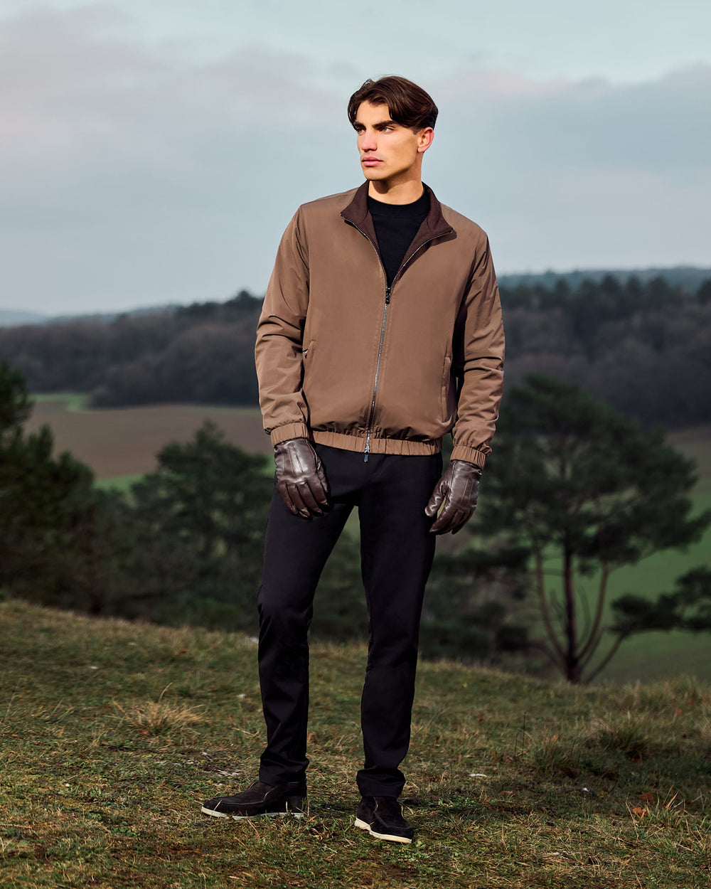 Man wearing a brown jacket and black pants standing in a field with trees and sky in the background