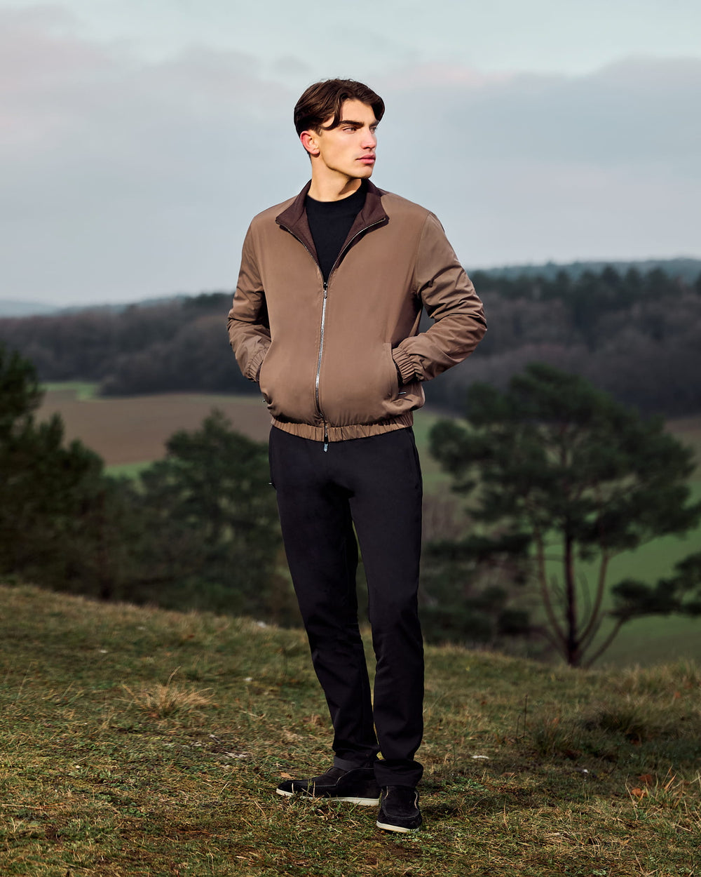 Man wearing a brown jacket standing in a natural landscape with trees and open fields.