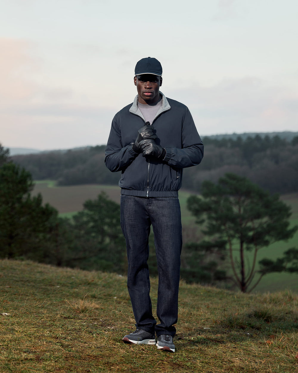 Man in outdoor setting wearing a dark jacket, cap, and gloves with a scenic background.