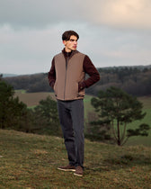 Man wearing a brown vest and dark pants standing in a field with trees and a cloudy sky.