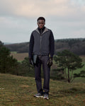 Man standing outdoors in a natural landscape wearing a dark jacket and pants.